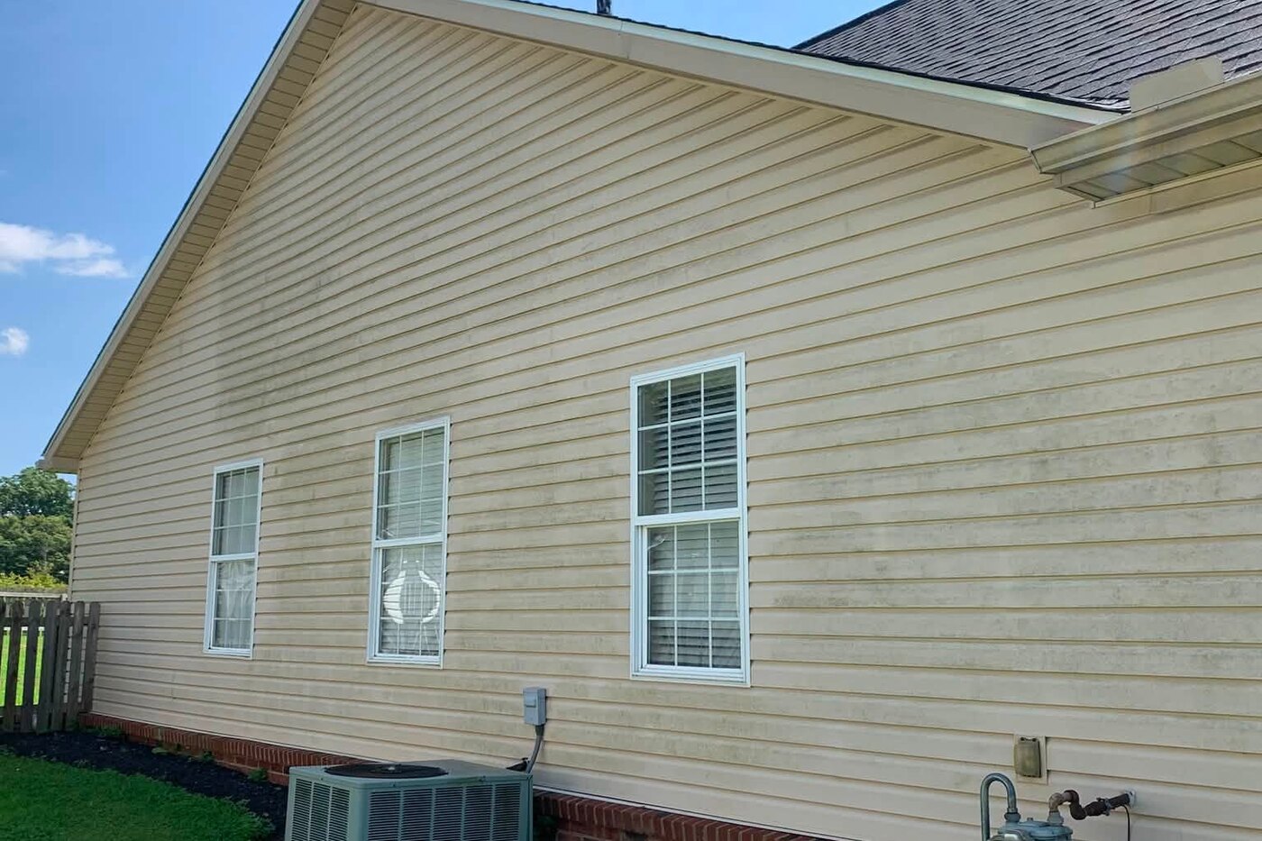 A gable end of a home with dirty cream siding above a brick skirt before washing